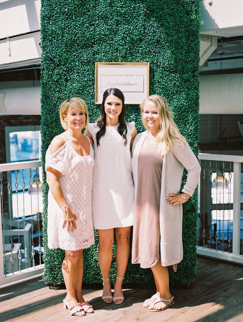Three women stand with hands on hip smiling in front of large greenery wall 