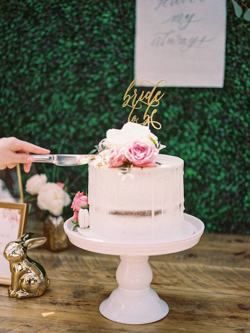 Hand with knife hovering over white naked cake with floral accents in front of greenery wall