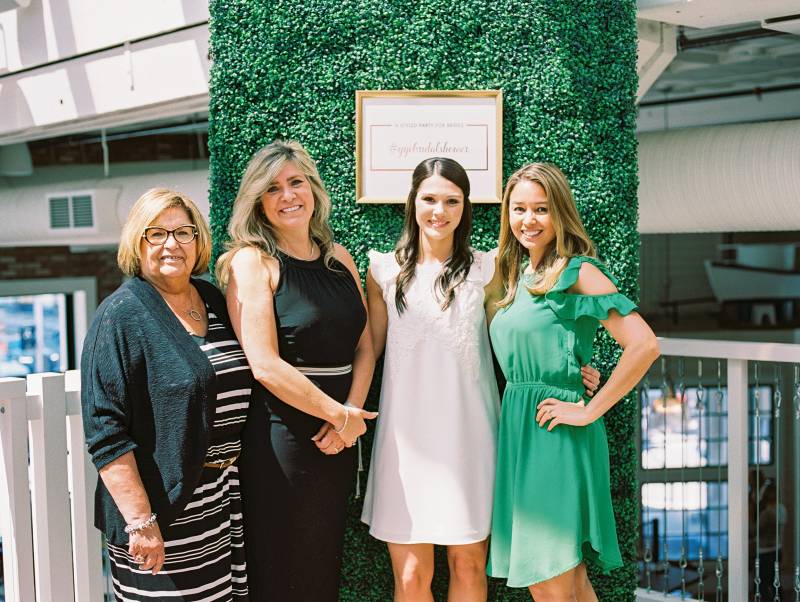Four women stand with hands crossed and on hip smiling in front of large greenery wall 