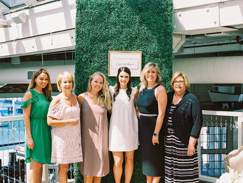 Six women stand smiling in front of large greenery wall 