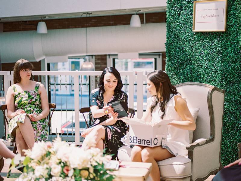 Three woman sitting watching gifts be opened by woman in white in front of large greenery wall 