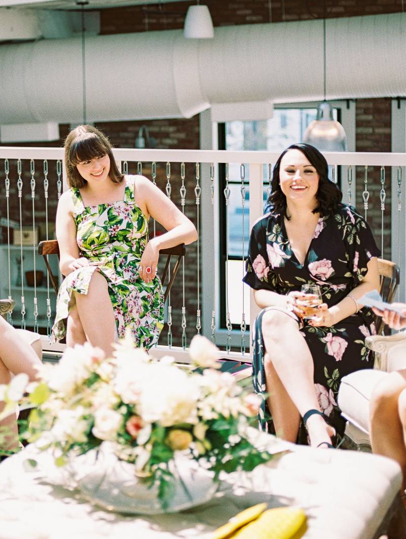 Two women in floral print dresses sitting and laughing behind white floral centerpiece 
