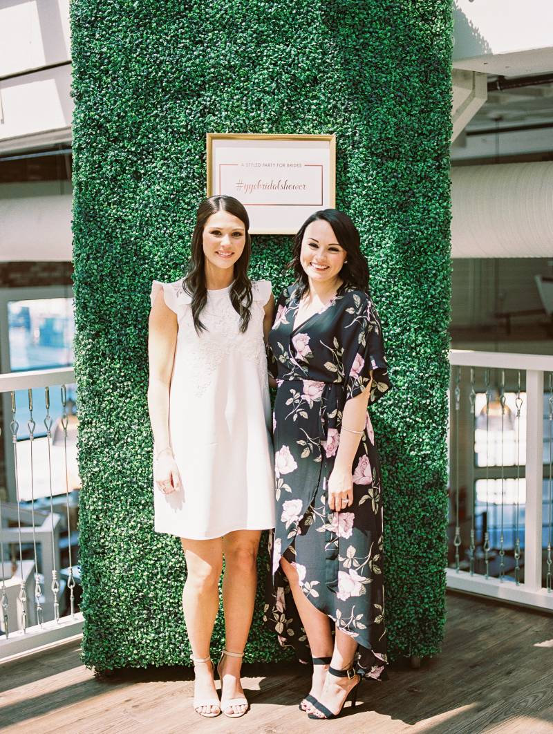 Two women in white and floral dresses stand together in front of large greenery wall 