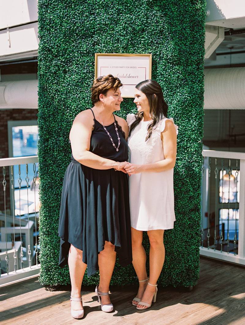 Two women one in black dress and one in white smiling at each other in front of large greenery wall 