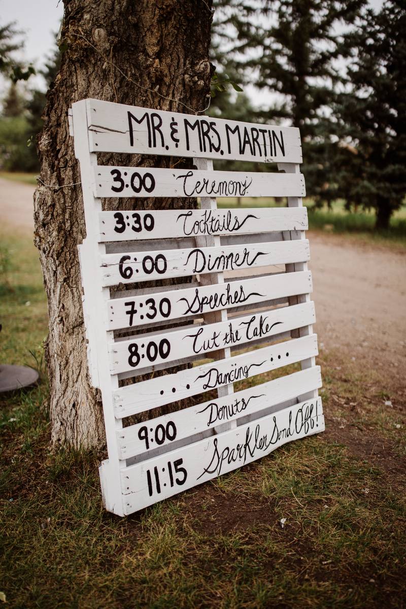 Wedding day timeline on white wood palate leaning against tree 