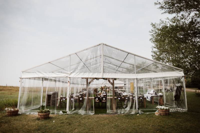 Wedding ceremony under clear tent with white and lavender floral accents and wooden wedding arch 
