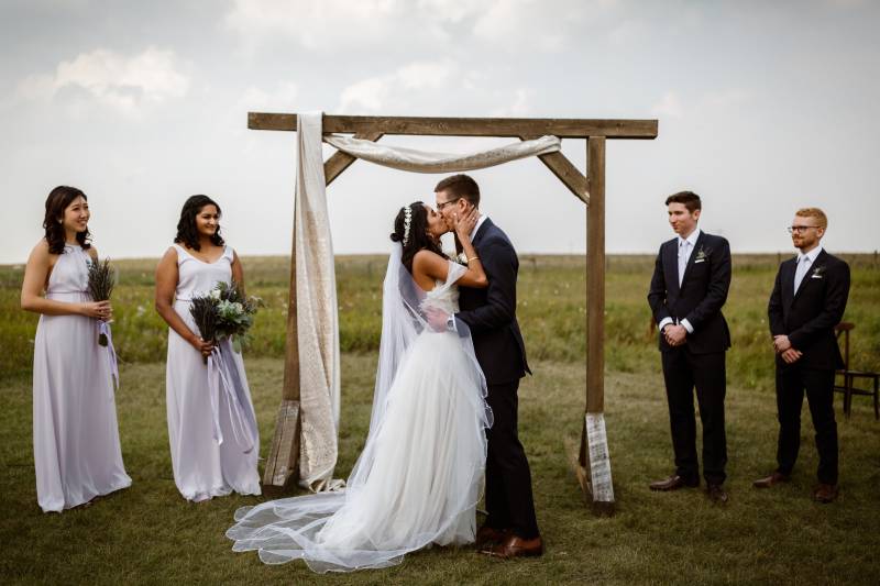 Bride and groom kiss in front of wedding arch with draping white tapestry on farm field 