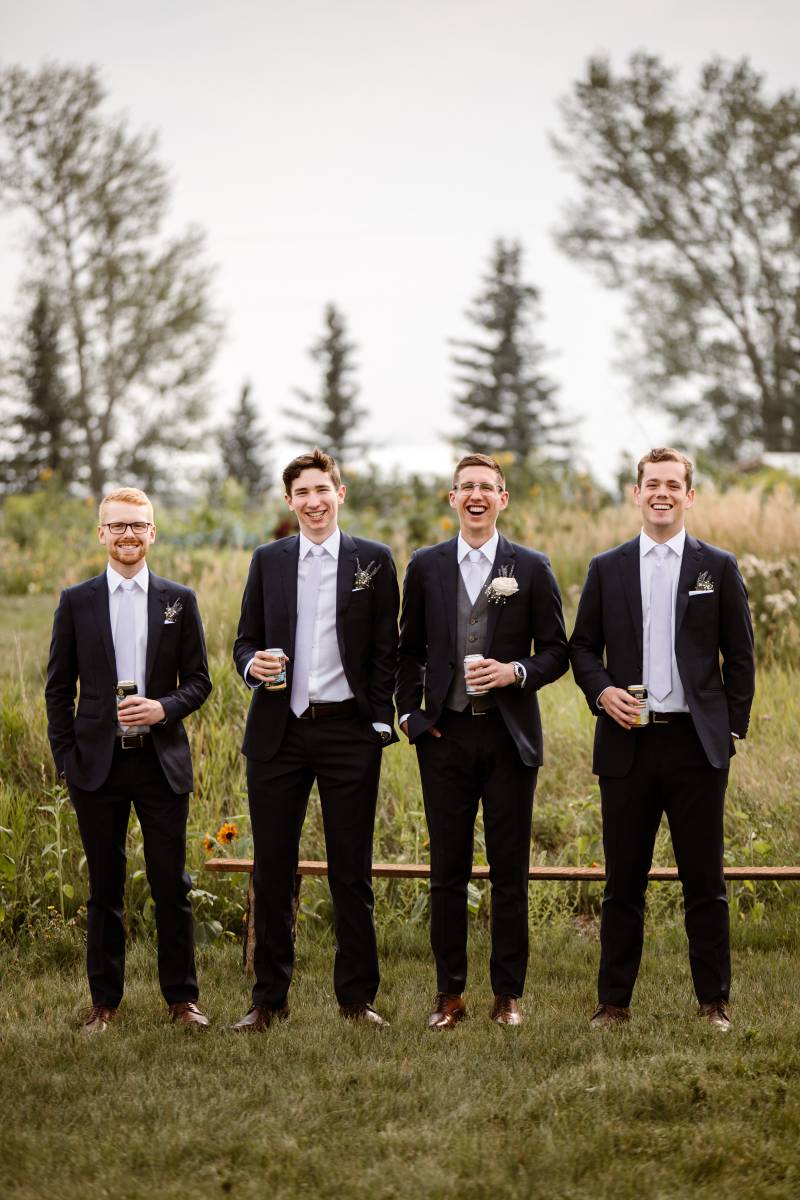 Groom and groomsman standing in sunflower patch laughing and drinking 