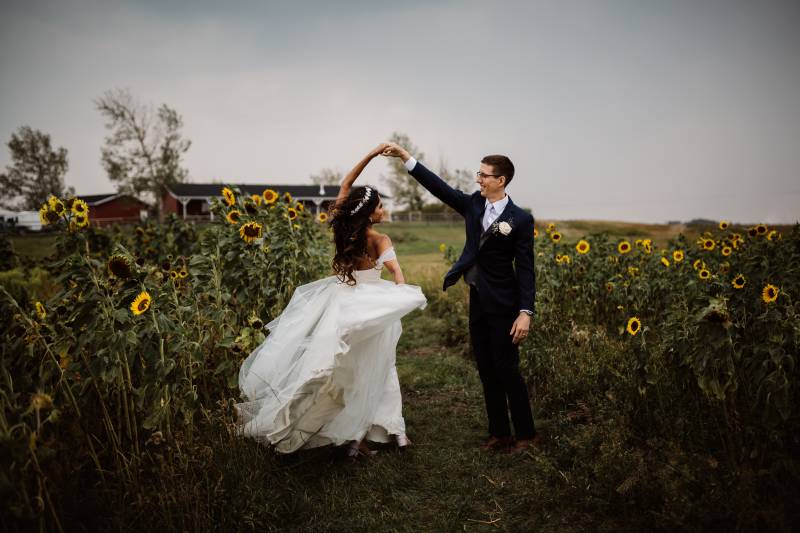 Groom spinning bride in sunflower patch wearing white off the shoulder dress and white flower crown