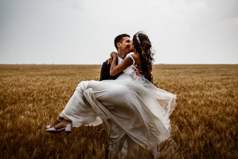 groom holding and kissing bride in wheat field with flowing white skirt