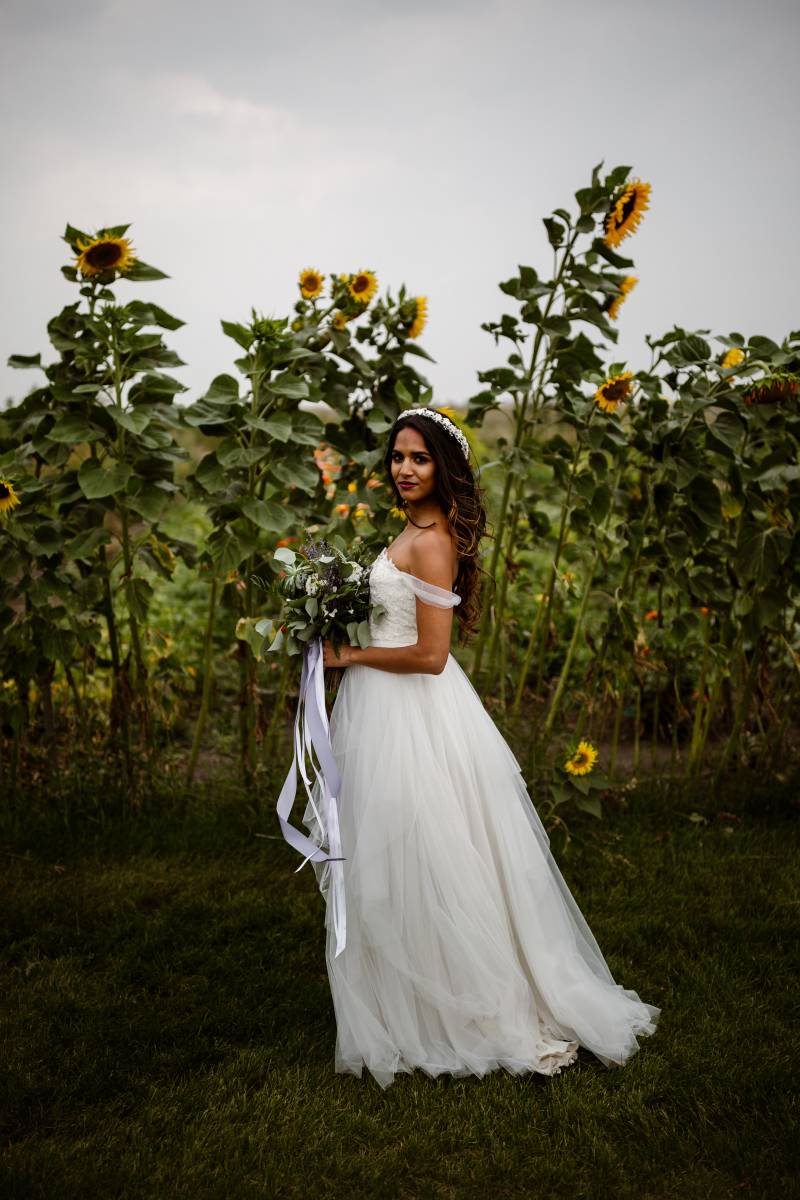 Bride standing in sunflower patch wearing white off the shoulder dress and lavender bouquet 