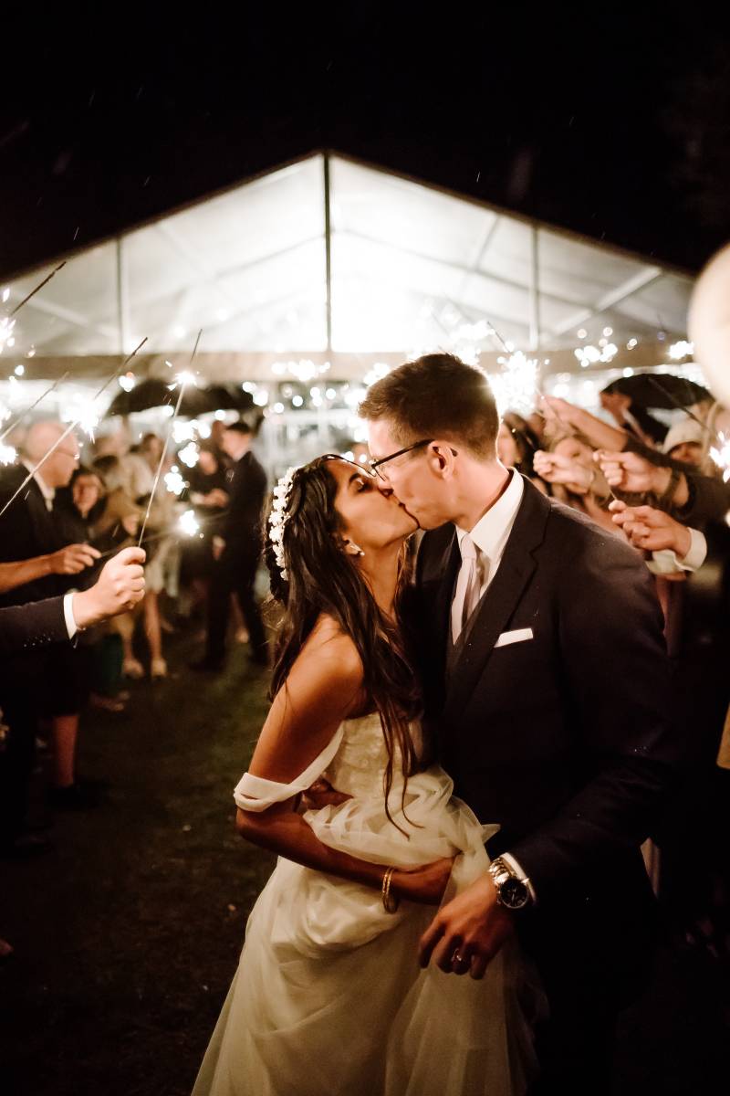 Bride and groom kiss in front of wedding reception tend as guests hold sparklers behind