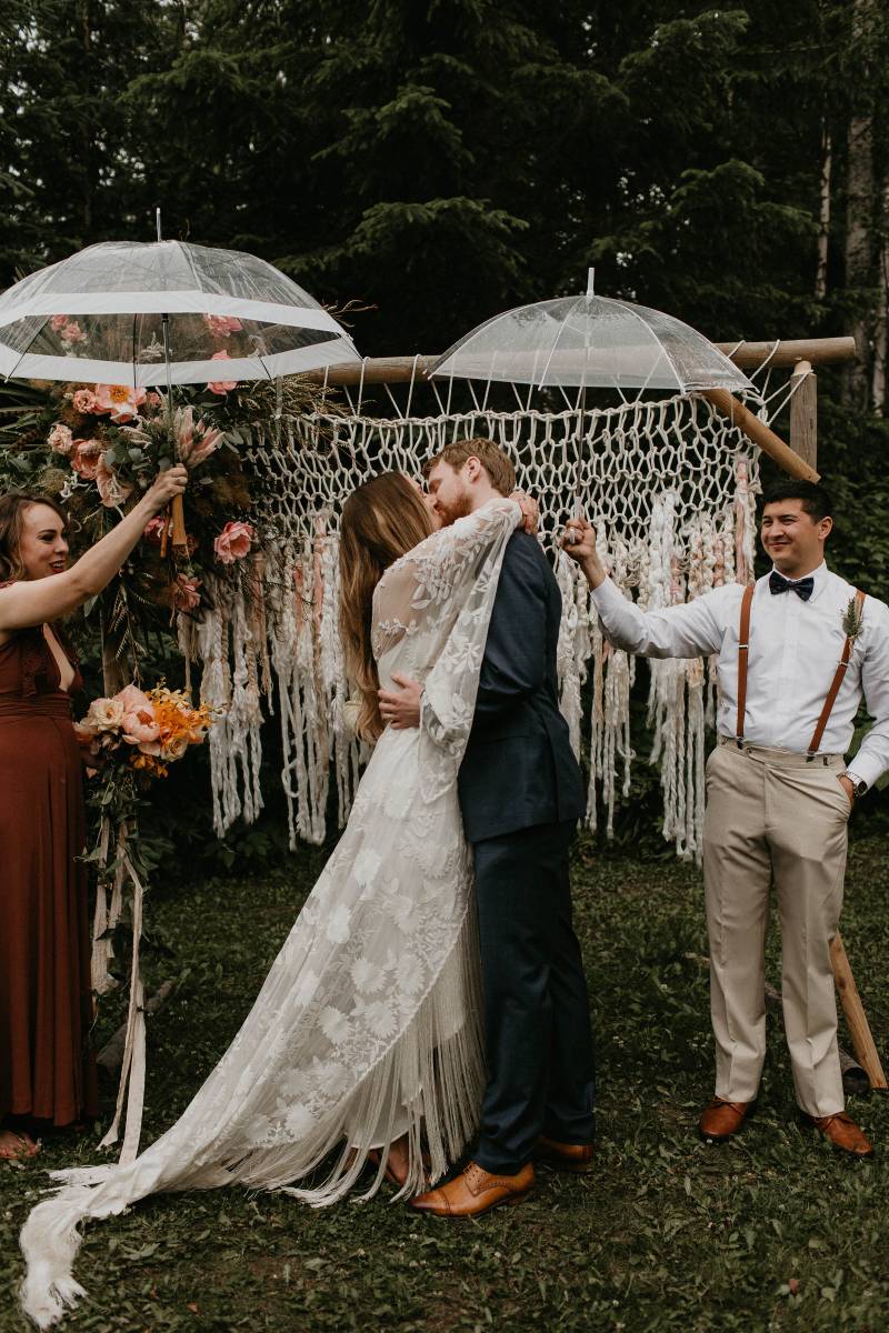Bride and groom kiss in front of macrame wedding arch while raining 