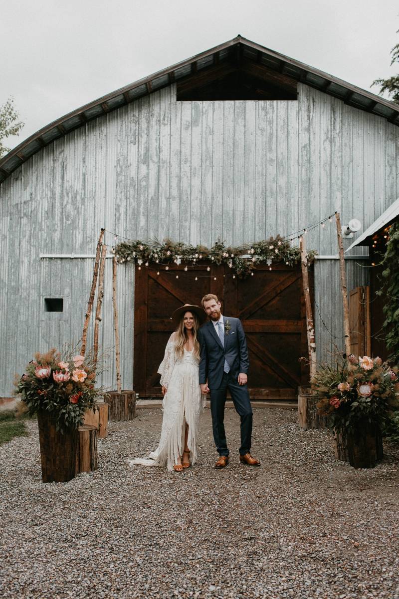 Bride and groom standing in front of barn and fairy light wedding arch 