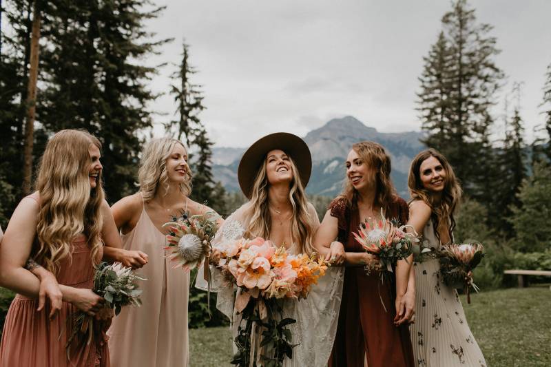 Bridesmaids in pink and blush dresses link arms in front of mountains 