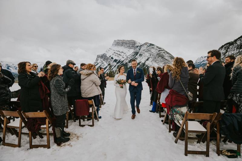 Bride and groom holding hands walk down snowy aisle while guests applaud 