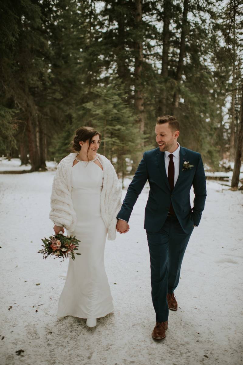 Bride and groom walk together in snowy forest holding hands 