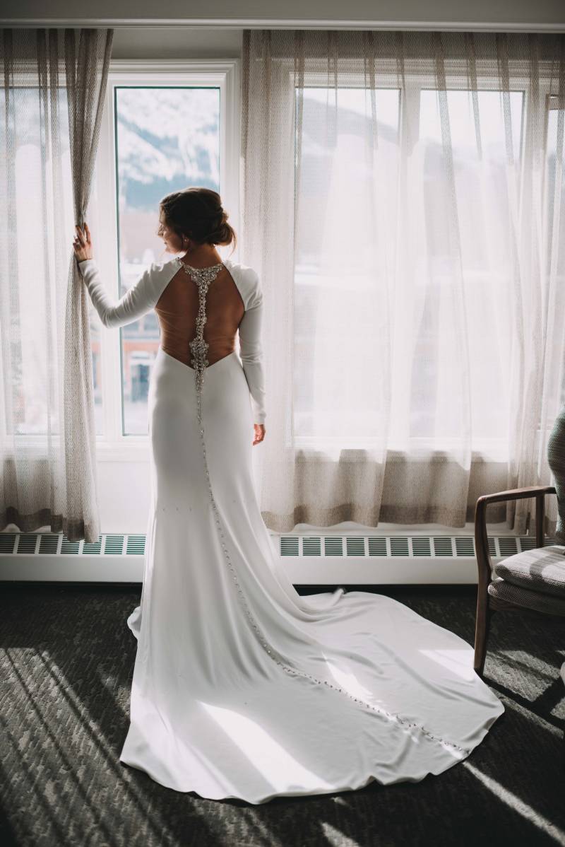 Bride in long open back dress stands at windowsill 