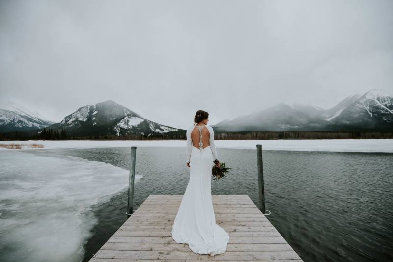 Bride stands in white dress at the end of frozen dock and mountains