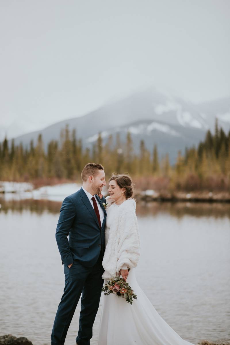 Bride and groom stand together in front of pond and snowy mountain 