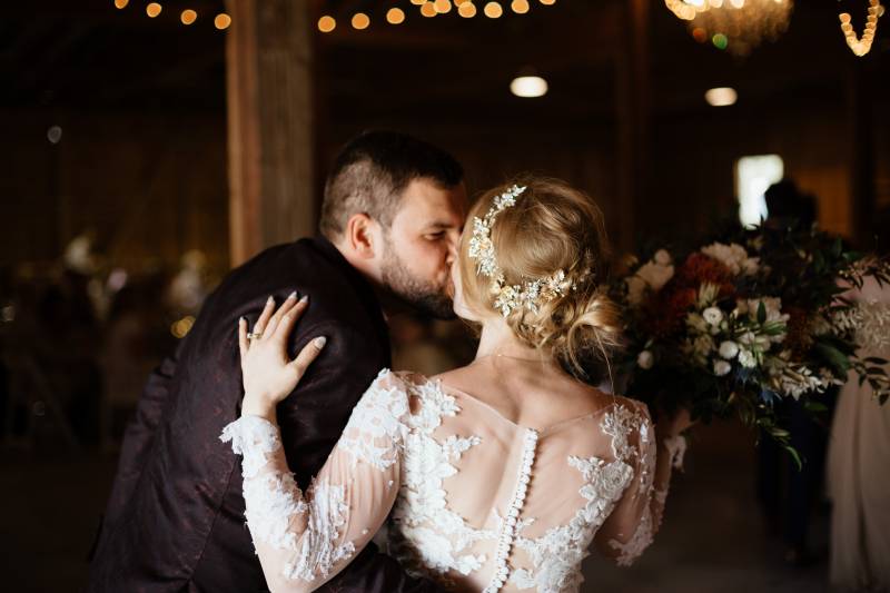 Bride and groom kiss in wooded barn wedding reception with floral arrangement background and fairy lights 