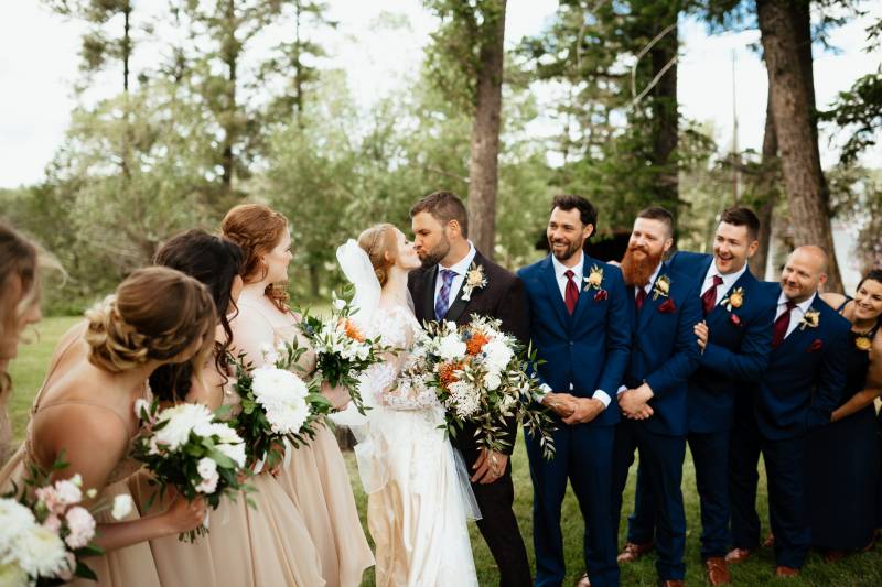 Bride and groom kiss as bridesmaids and groomsmen smile and lean 