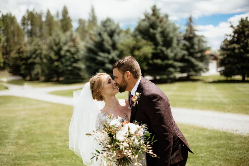 Bride wearing white veil and white bouquet kissing groom with forest backdrop