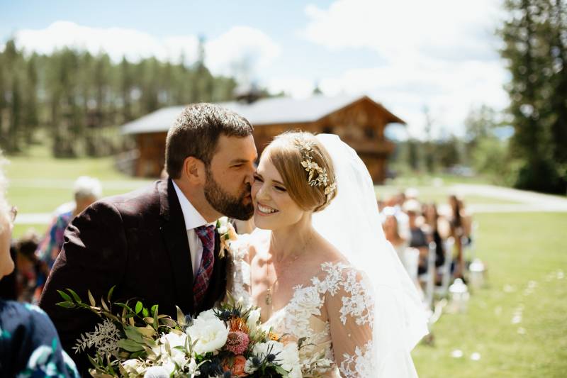 Groom kissing bride in white lace dress and veil on the cheek with cabin and forest backdrop