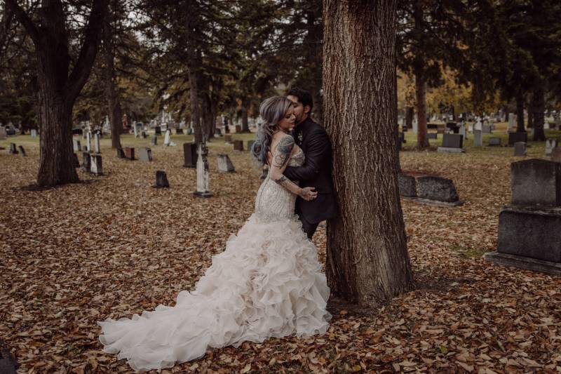 Bride leaning against groom leaning on tree amidst fall leaves in cemetery 