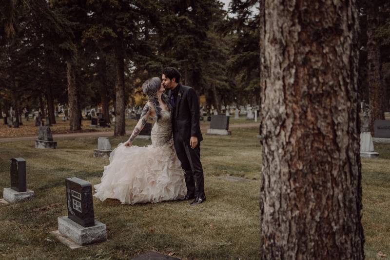 Bride and groom kiss embraced in cemetery while holding white dress behind 