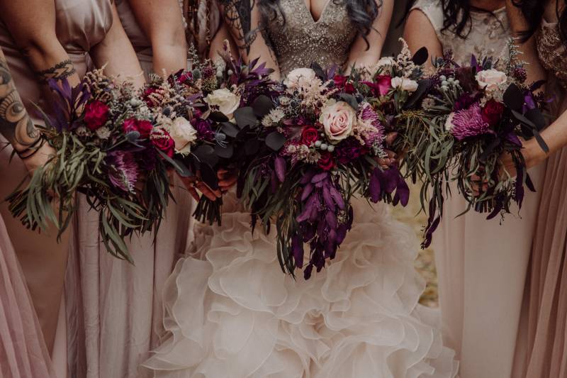 Bride and bridesmaids hold out purple white pink bouquets together in line 