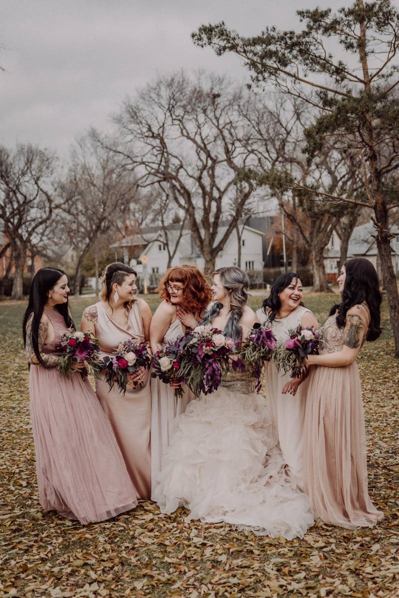 Bride and bridesmaids stand holding burgundy and white bouquets in fall leafy field 