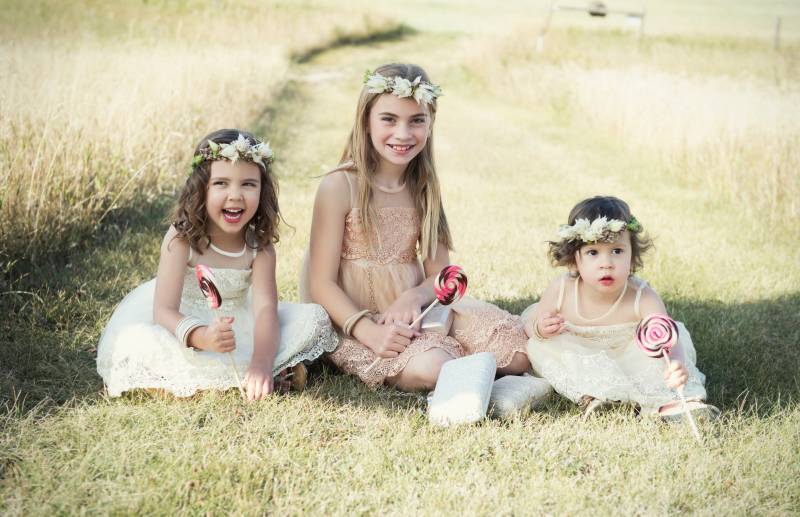 Three flower girls sit in a row wearing white flower crowns and blush dresses holding pink white and brown lollipops on grassy pathway 