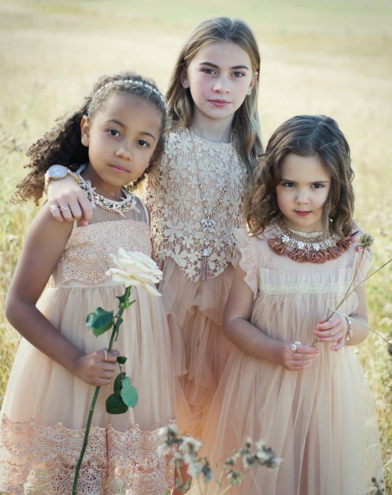 Three flower girls in blush lace dresses stand holding white flowers in grassy field 