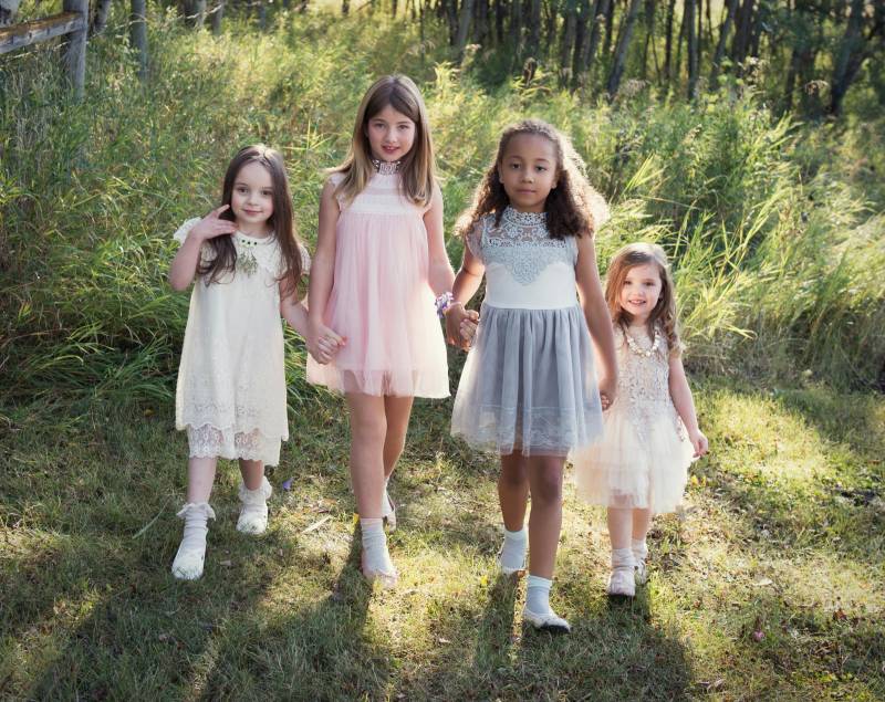 four flower girls walk holding hands wearing pale lace dresses during sunset in grassy field 