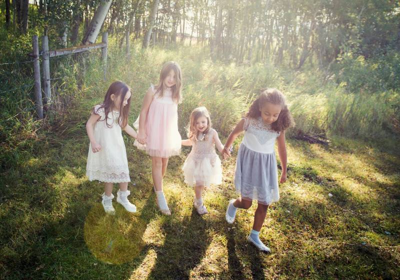 four flower girls walk holding hands wearing pale lace dresses during sunset in grassy field 