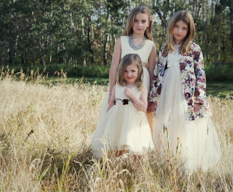 Three flower girls stand wearing pale yellow dresses and floral overcoat in brown grass field 