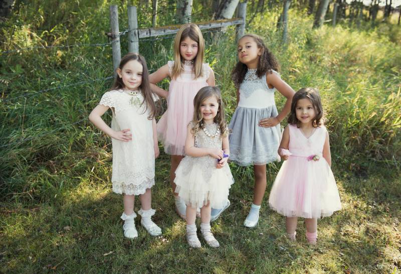 Five flower girls stand smiling with hands on hips in green grass forest