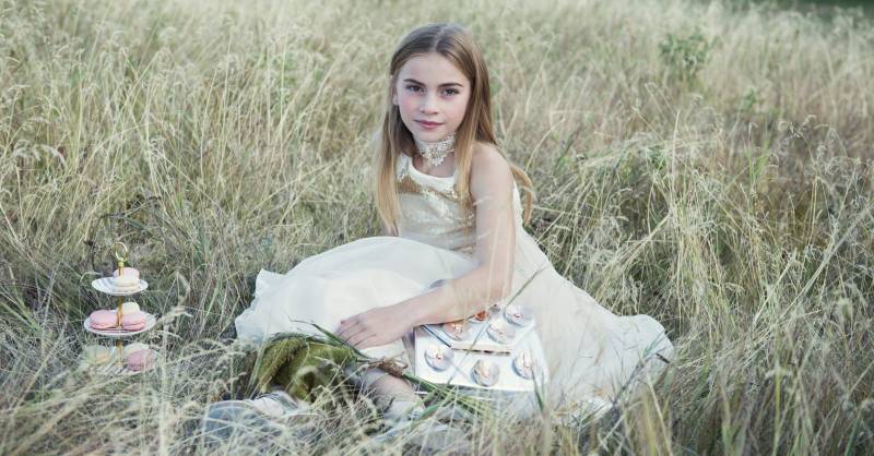 Flowergirl sits in white and gold lace dress with silver clutch beside display of macaroons in grassy field 