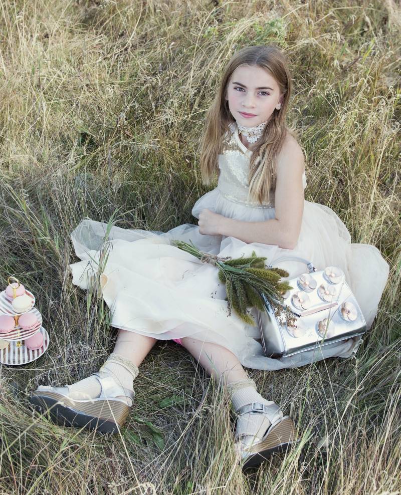 Flower girl in white and gold dress sits with silver purse and green bouquet beside plate of macaroons in grassy field 