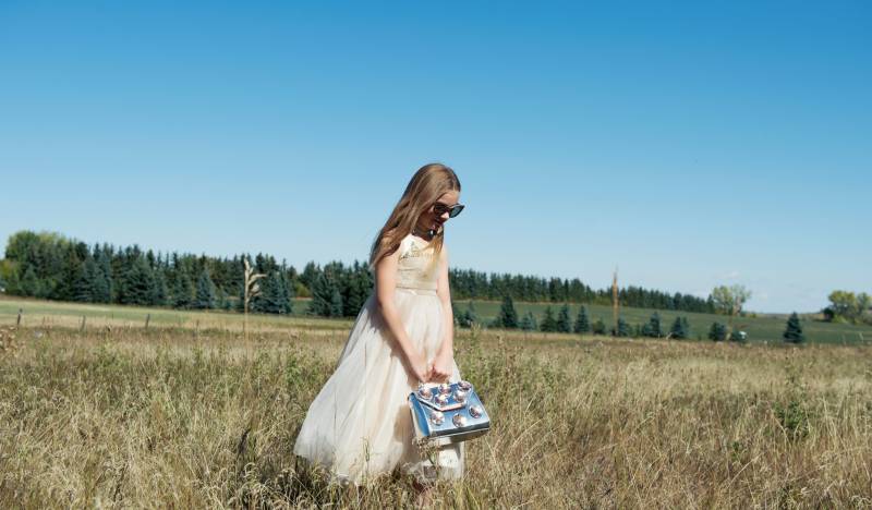 Flower girl in white and gold dress and sunglasses holding silver clutch in grassy field 