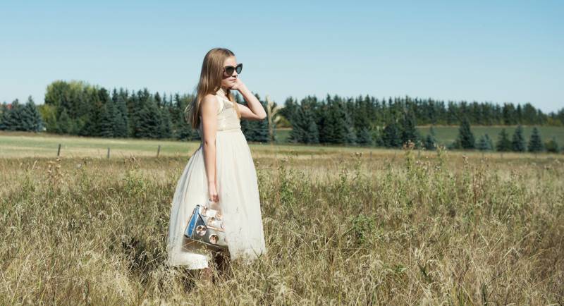 Flower girl in white and gold dress holding silver clutch adjusts sunglasses in grassy field 
