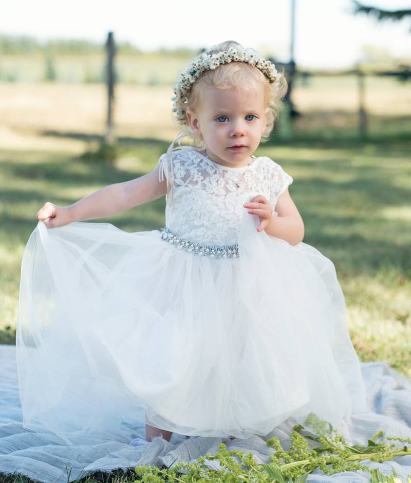 Flower girl in white lace dress and white flower crown walks on white fabric in grassy field 