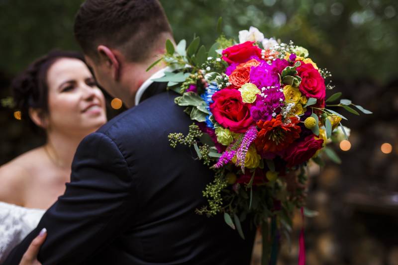 Bride holds multicolored bouquet behind groom while looking into eyes