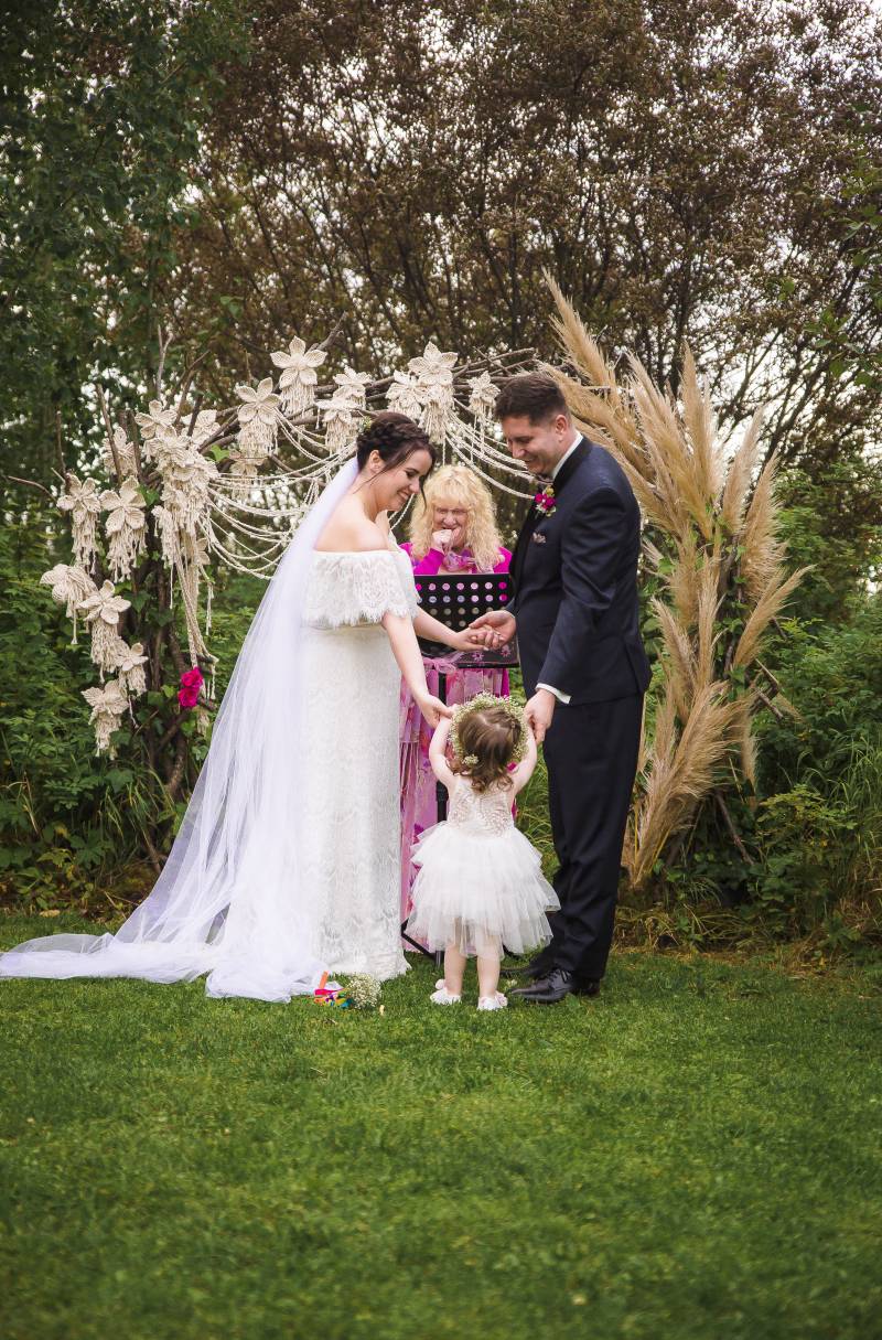 Small child hands rings to bride and groom standing in front of macramé wedding arch 