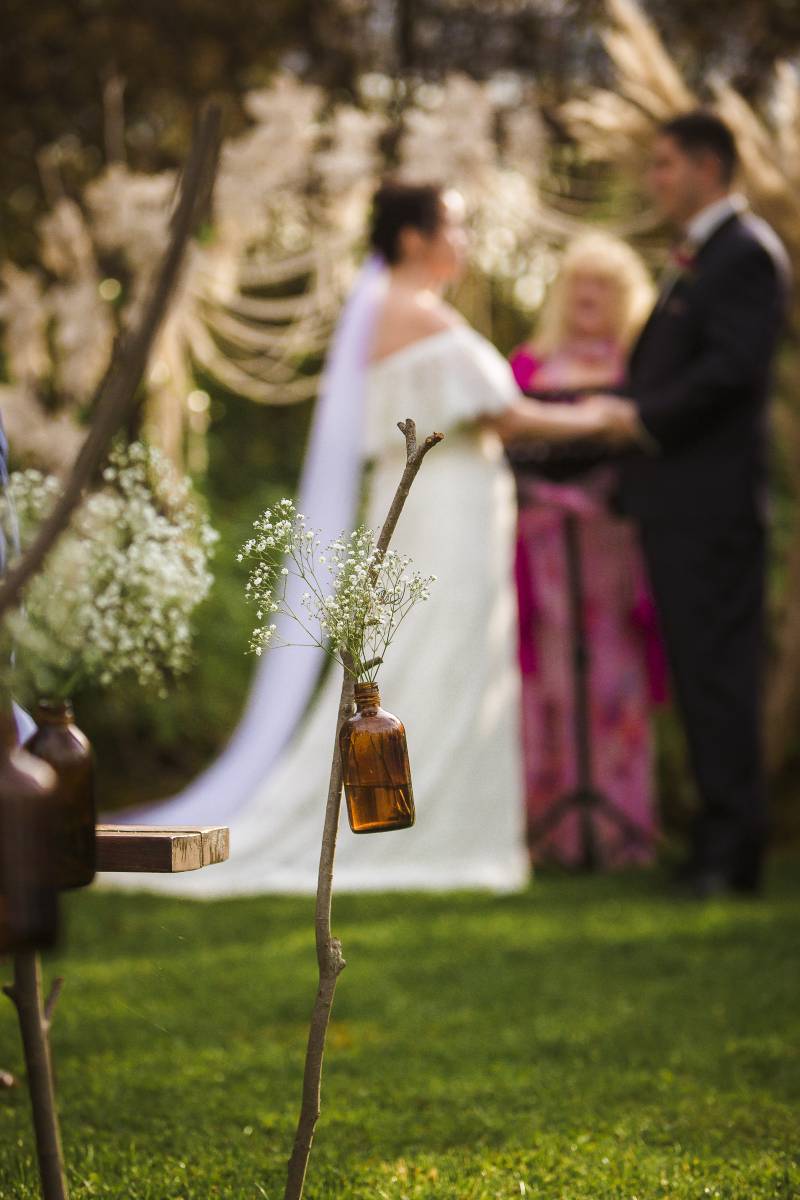 Bride and groom stand together behind stick with bottle holding white flower
