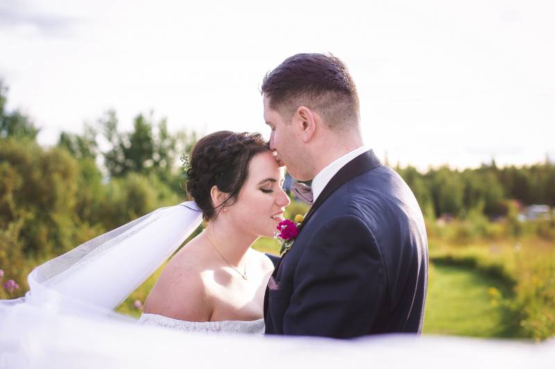 Groom kisses brides forehead in grassy meadow 