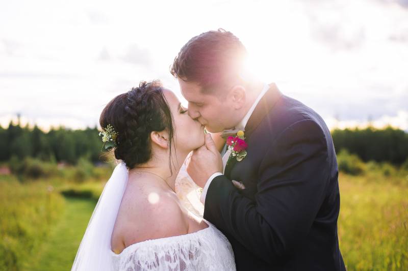 Bride and groom kiss while sun sets on grassy meadow