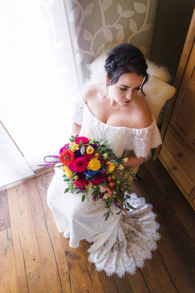 Bride sitting in white lace dress holding lush red yellow and purple bouquet