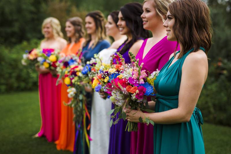Bridesmaids stand in line wearing multicolored dresses and bouquets